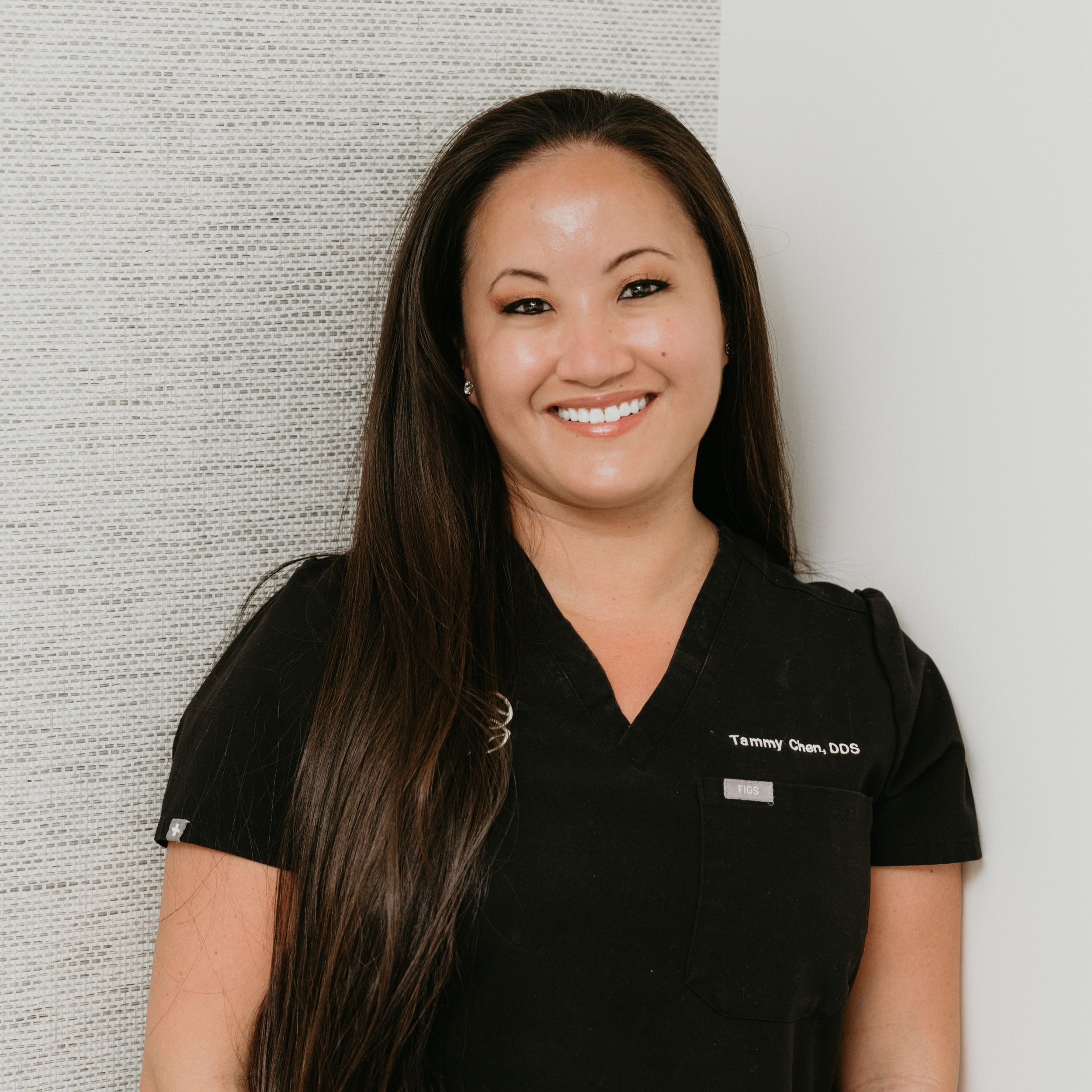 The image features a smiling woman wearing a white lab coat with name tags, standing against a textured wall with a light-colored background.