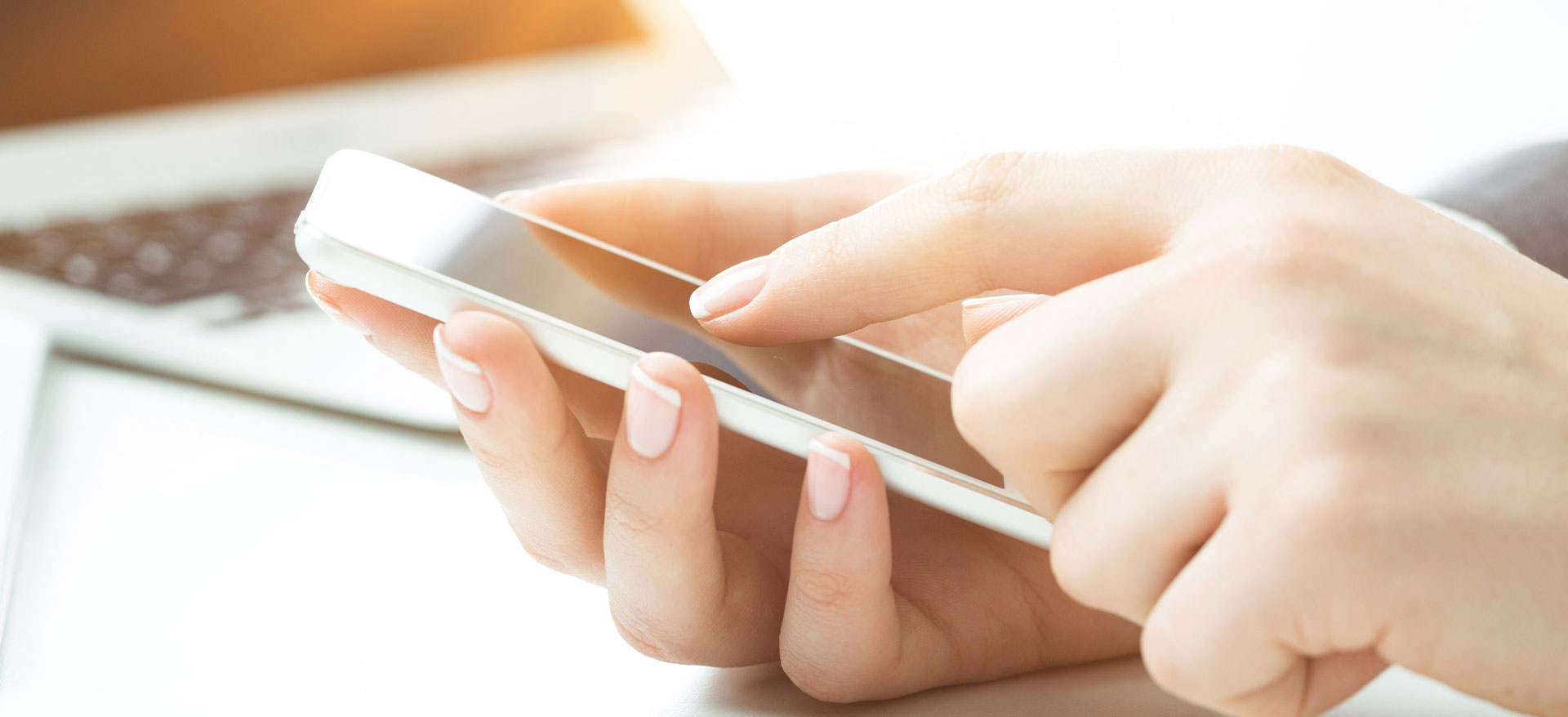 A person s hand holding a smartphone with a blurred background featuring a laptop and a desk.