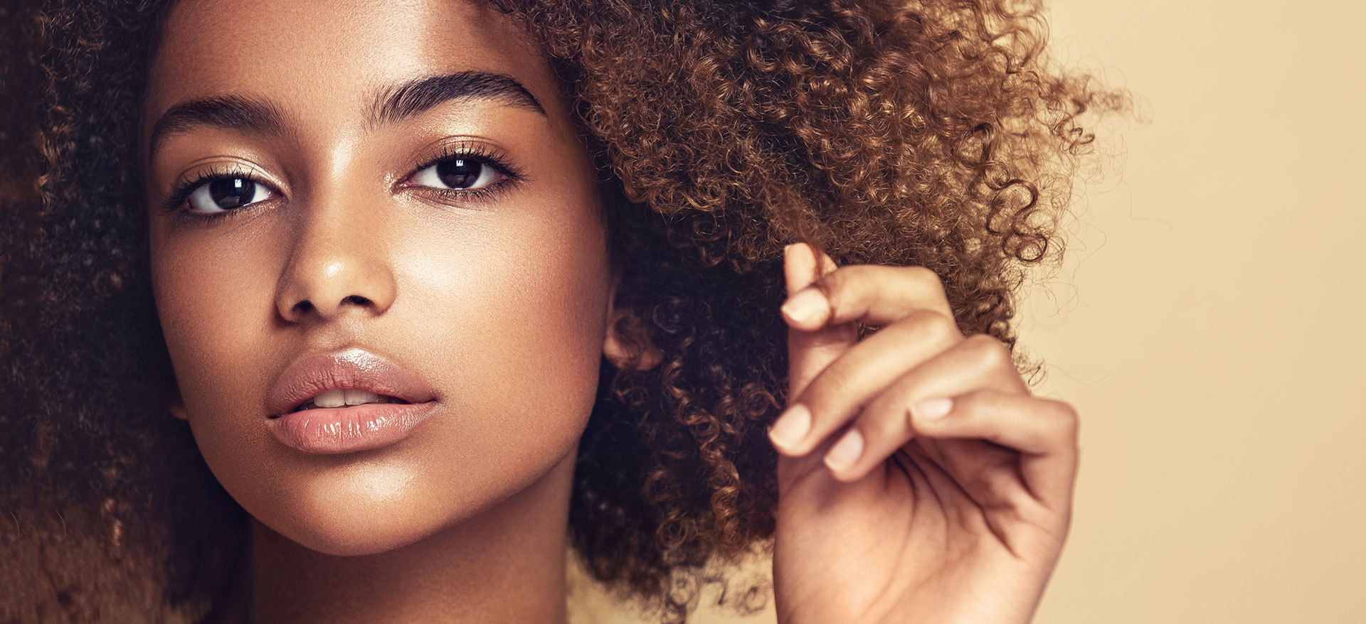 A woman with curly hair and makeup, wearing a yellow top, set against a neutral background.