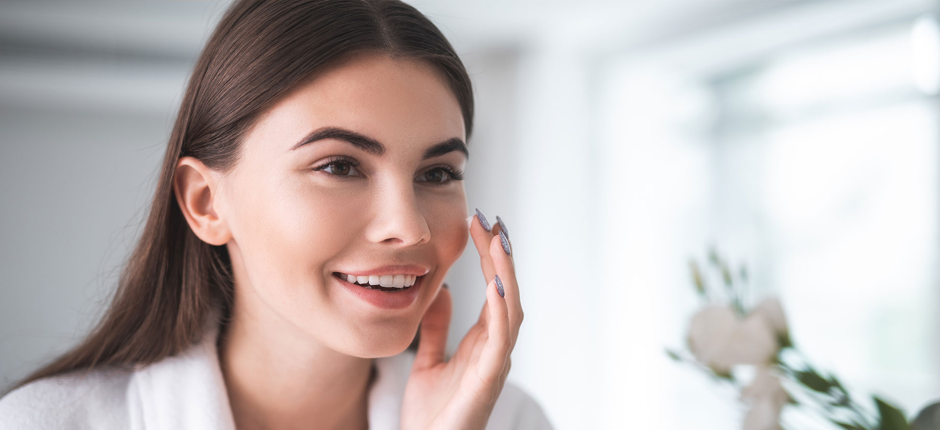 A woman with a neutral expression looking at her face in a mirror while applying makeup.