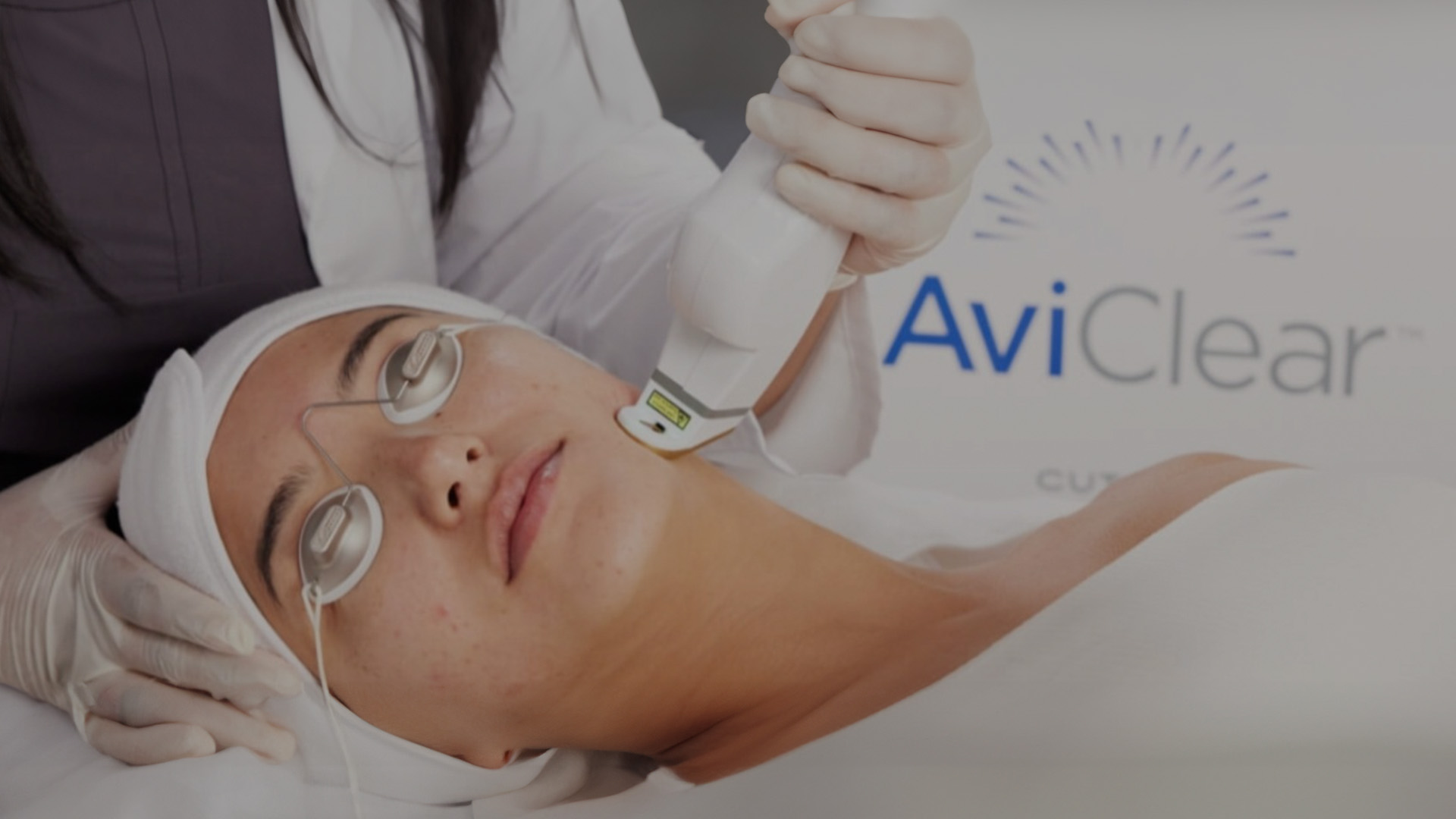 A person receiving a beauty treatment with a device on their face while wearing protective eyewear, attended by a professional in a medical setting.