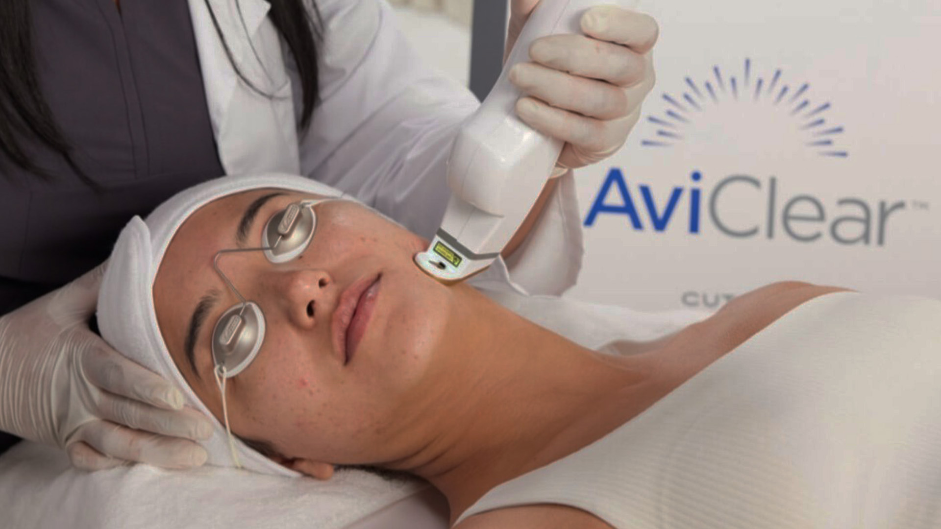 A woman receiving a facial treatment using a machine, with an attendant applying the device on her face, all set against a backdrop that includes an AvivClear sign.