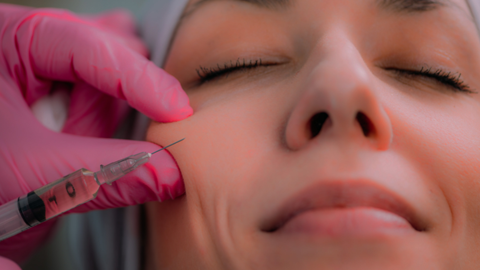 A person receiving a cosmetic procedure, with a syringe being injected into their face by a professional wearing medical gloves and a pink mask.