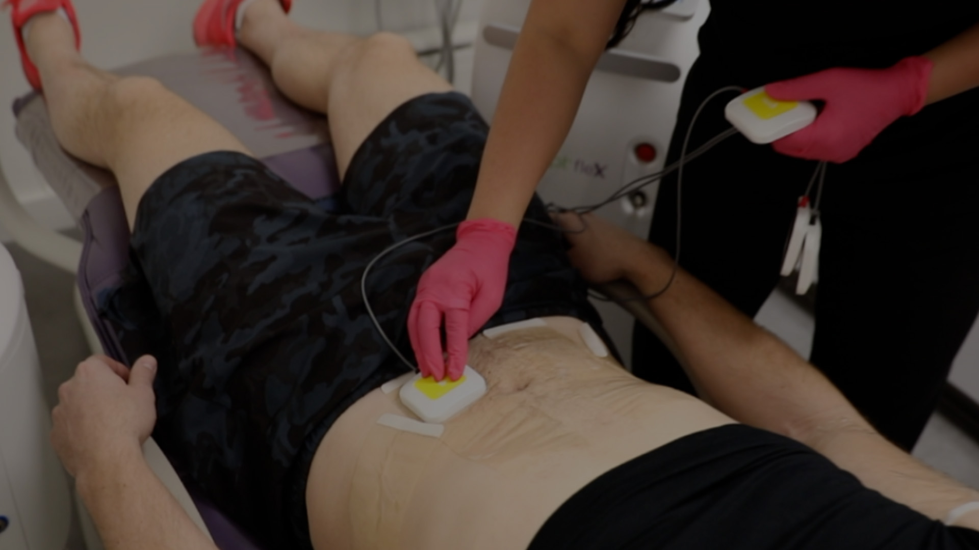 The image shows a person lying on an examination table with a medical device attached to their abdomen, being monitored by a healthcare professional wearing gloves and a face mask, indicating a clinical setting.