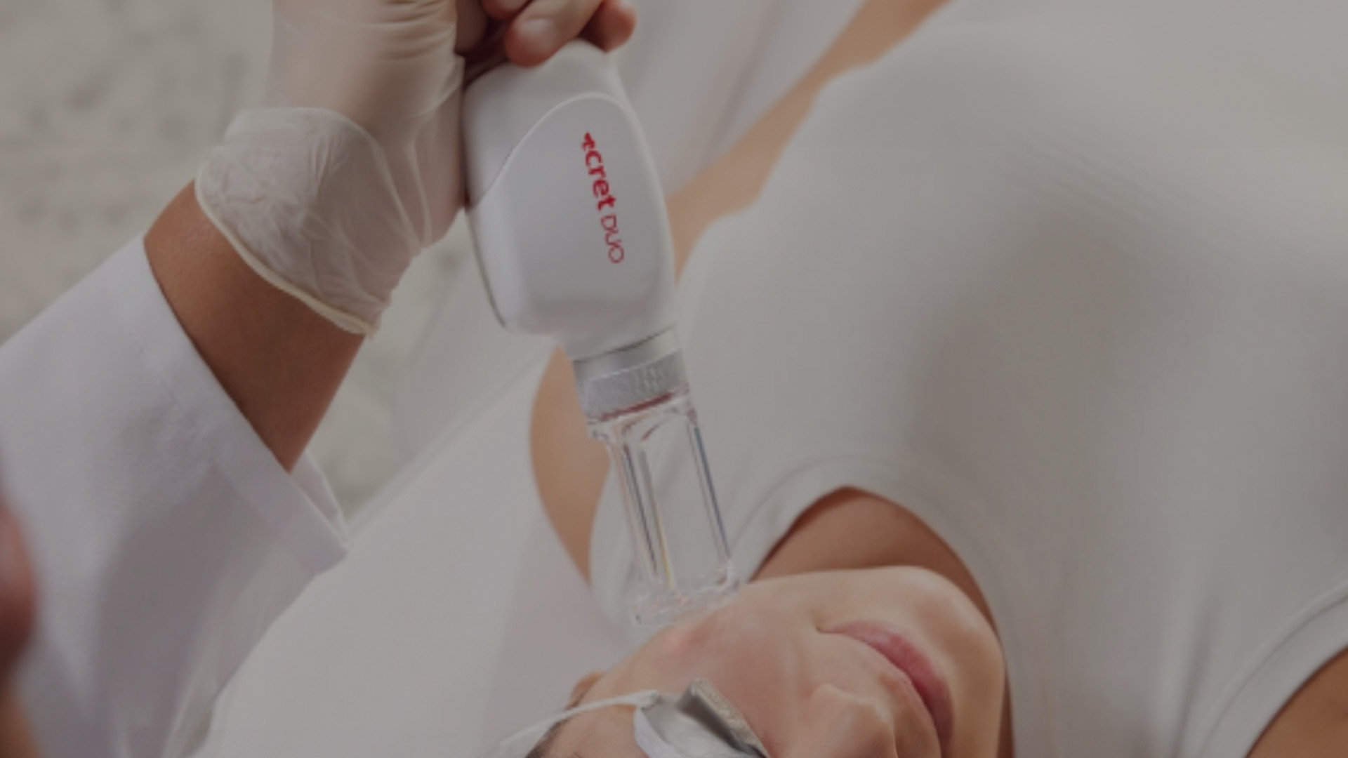 A person receiving a cosmetic treatment with a handheld device, while another person, presumably a technician, monitors the process.