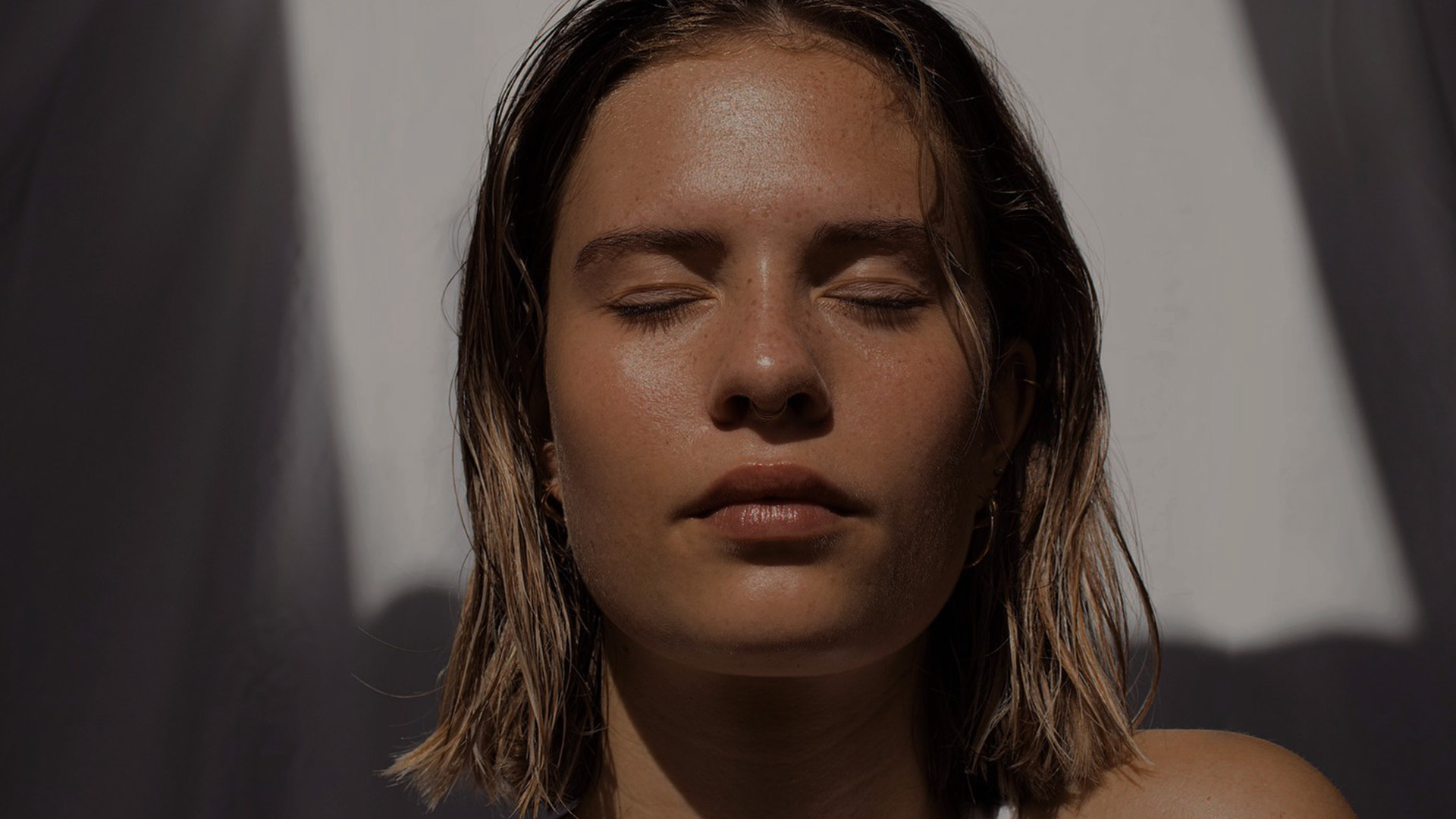 A young woman with short hair, wearing a white sleeveless top, has her eyes closed and appears to be meditating or resting with a serene expression.
