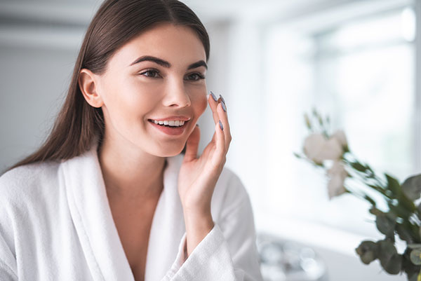 A woman applying skincare products while standing in front of a mirror in a well-lit room with plants in the background.