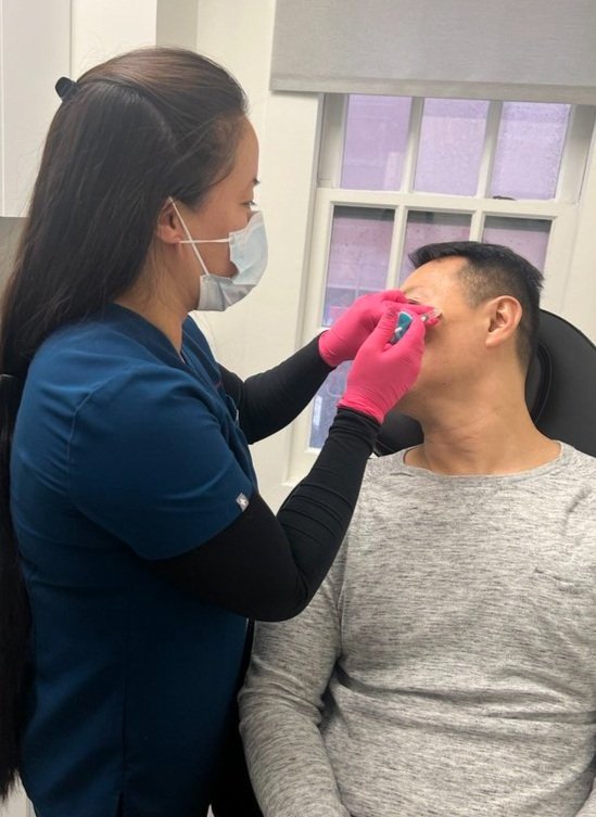 A woman wearing a surgical mask and gloves is applying a blue device to a man s face while he sits in a dental chair.