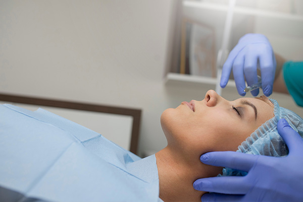 The image shows a woman lying on a bed with her eyes closed, receiving medical attention from a healthcare professional who appears to be performing a procedure using a device that may be a surgical tool or a diagnostic instrument.